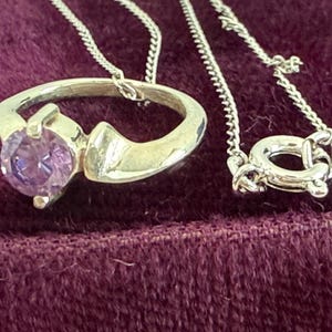 May include: A silver pendant necklace featuring a ring-shaped design with a large, round, purple gemstone. The necklace chain and clasp are also silver. The jewelry is displayed on a dark purple velvet surface.