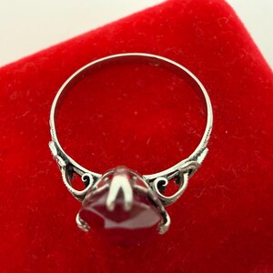 May include: A silver ring with a large, faceted, red gemstone. The ring has ornate detailing on the band and around the gemstone setting. The ring is displayed on a red velvet surface.
