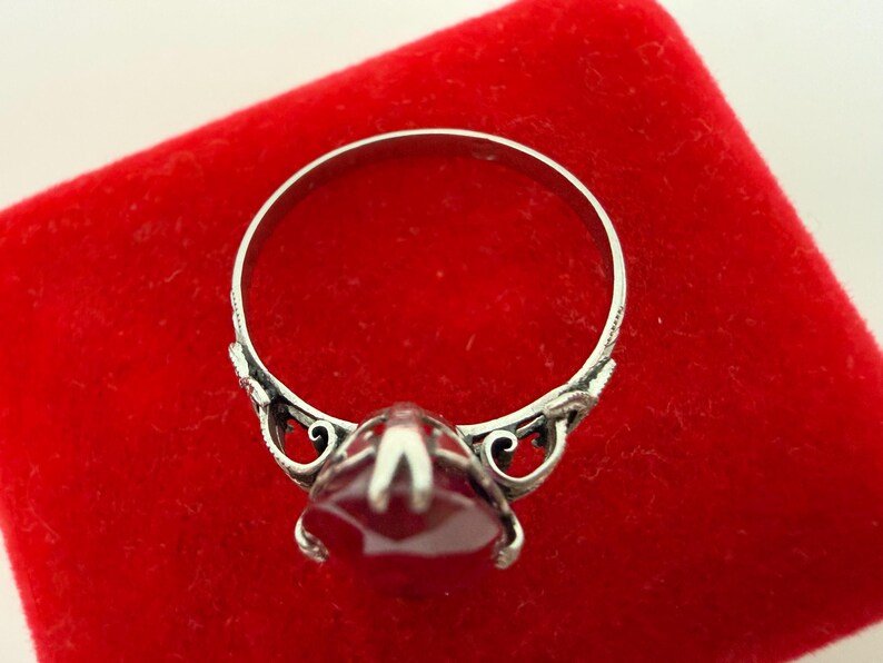 May include: A silver ring with a large, faceted, red gemstone. The ring has ornate detailing on the band and around the gemstone setting. The ring is displayed on a red velvet surface.
