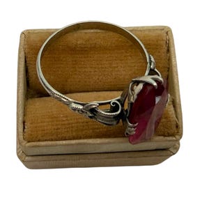 May include: A silver ring with a large, faceted, deep red gemstone. The ring has an ornate, floral design on the band and setting. The ring is displayed on a small, tan, velvet-lined box.