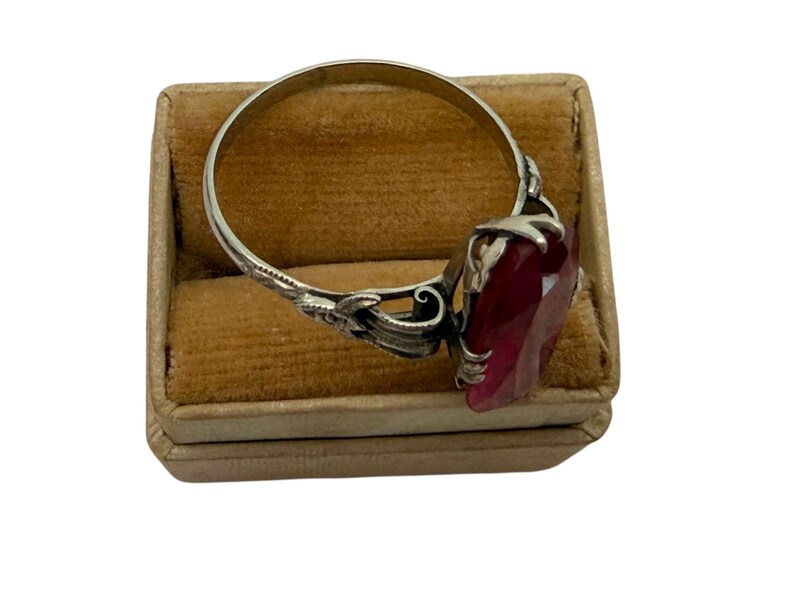 May include: A silver ring with a large, faceted, deep red gemstone. The ring has an ornate, floral design on the band and setting. The ring is displayed on a small, tan, velvet-lined box.