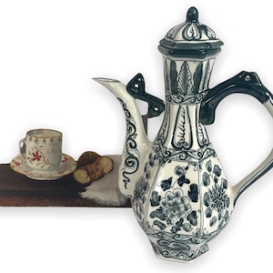 May include: A white and blue ceramic coffee pot with a floral design, a matching cup and saucer, and two pastries. The coffee pot has a tall, ornate shape with a lid and handle. The cup and saucer have a floral pattern.