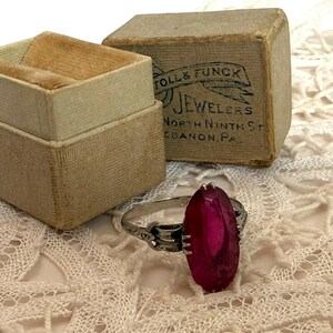 May include: A vintage silver ring with a large, oval, magenta-colored stone. The ring sits on a white lace surface next to an open, beige ring box with a velvet interior and a small, rectangular box with the text "TOLL & FUNCK JEWELERS".