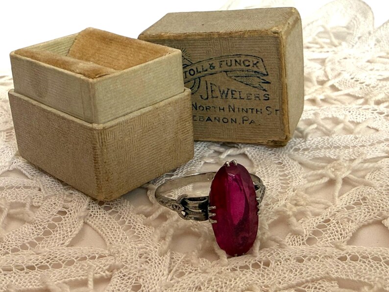 May include: A vintage silver ring with a large, oval, magenta-colored stone. The ring sits on a white lace surface next to an open, beige ring box with a velvet interior and a small, rectangular box with the text "TOLL & FUNCK JEWELERS".