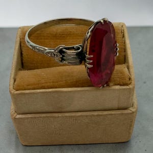 May include: A silver ring with an oval, faceted, red gemstone. The ring is set in a decorative setting and rests on a small, tan velvet jewelry box. The ring's band has an ornate design near the gemstone.