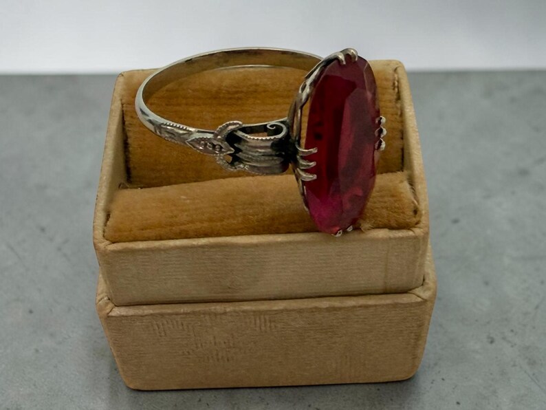 May include: A silver ring with an oval, faceted, red gemstone. The ring is set in a decorative setting and rests on a small, tan velvet jewelry box. The ring's band has an ornate design near the gemstone.