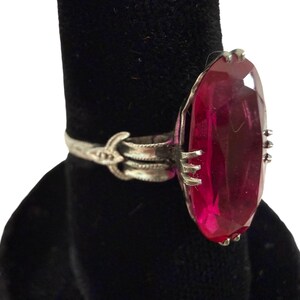 May include: A silver ring featuring a large, oval-cut, deep red gemstone. The ring has an ornate band with decorative detailing and is displayed against a black background. The gemstone is the focal point, catching the light.