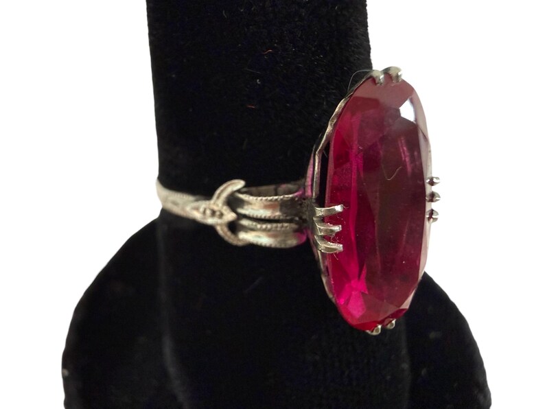May include: A silver ring featuring a large, oval-cut, deep red gemstone. The ring has an ornate band with decorative detailing and is displayed against a black background. The gemstone is the focal point, catching the light.