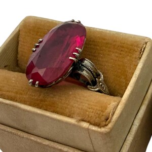 May include: A vintage ring with a large, faceted, oval-shaped, deep red gemstone. The ring is set in a silver-toned metal with ornate detailing and is displayed in a small, tan velvet-lined jewelry box. The ring is a statement piece.