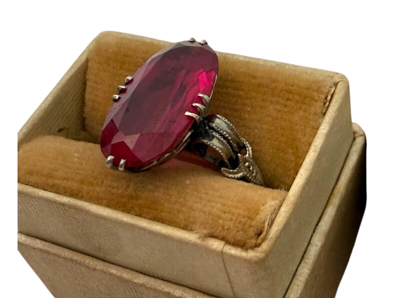 May include: A vintage ring with a large, faceted, oval-shaped, deep red gemstone. The ring is set in a silver-toned metal with ornate detailing and is displayed in a small, tan velvet-lined jewelry box. The ring is a statement piece.