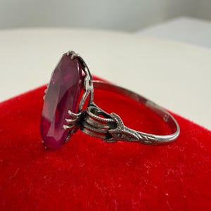May include: A silver ring with a large, oval-cut, deep red gemstone. The ring has an ornate, detailed setting with a floral design on the band. The ring is displayed on a red velvet surface.