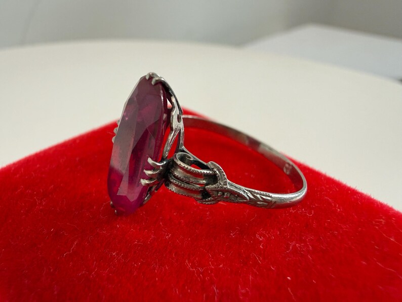 May include: A silver ring with a large, oval-cut, deep red gemstone. The ring has an ornate, detailed setting with a floral design on the band. The ring is displayed on a red velvet surface.