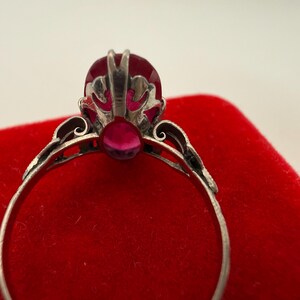 May include: Close-up of a silver ring with a large, oval-shaped, red gemstone as the centerpiece. The ring has intricate filigree detailing around the gemstone and along the band. The ring is set against a red velvet surface.