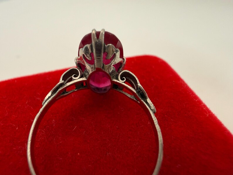 May include: Close-up of a silver ring with a large, oval-shaped, red gemstone as the centerpiece. The ring has intricate filigree detailing around the gemstone and along the band. The ring is set against a red velvet surface.