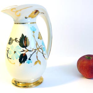 May include: Cream-colored ceramic pitcher with a gold rim and handle, adorned with blue floral and gold leaf designs. A red apple sits beside the pitcher. Decorative kitchenware.