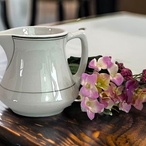 May include: A white ceramic pitcher with a black trim and handle. The pitcher is next to a bouquet of pink and purple flowers. The pitcher is on a dark wooden surface.