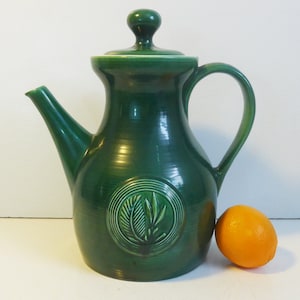 May include: A dark green ceramic pitcher with a lid and handle. The pitcher has a spout and a circular emblem with a leaf design. An orange is next to the pitcher.