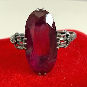 May include: A silver ring with a large, oval, faceted, deep red gemstone. The ring's band features intricate, stylized claw-like designs on either side of the gemstone, adding a unique and gothic aesthetic. The ring is displayed on a red surface.