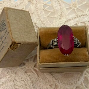 May include: An antique ring with a large, oval, ruby-colored stone set in a silver band. The ring is displayed in a small, vintage ring box next to a worn cardboard box with text that reads "Sole & Jungk Jewelers, North Ninth St, Lebanon, Pa."