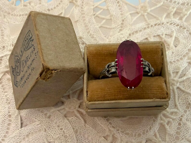 May include: An antique ring with a large, oval, ruby-colored stone set in a silver band. The ring is displayed in a small, vintage ring box next to a worn cardboard box with text that reads "Sole & Jungk Jewelers, North Ninth St, Lebanon, Pa."