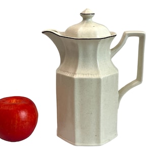 May include: A cream-colored, lidded pitcher with a geometric design and a black trim. The pitcher has a handle and a spout. A red apple is next to the pitcher. The pitcher is likely for serving beverages.