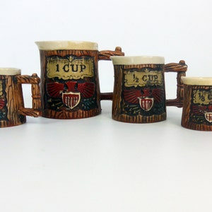 May include: Set of four ceramic measuring cups in the shape of wooden mugs. Each mug has a cream-colored rim and a handle. The mugs are decorated with a red eagle emblem and the measurements: 1 cup, 1/2 cup, and 1/3 cup.