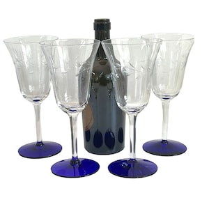 May include: Four clear wine glasses with etched leaf designs, each with a long stem and a deep blue base. A dark wine bottle stands behind the glasses. The glasses are arranged in a semi-circle.