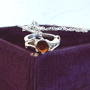 Peut inclure: Une bague en argent ornée d'une grosse pierre précieuse ronde de couleur ambre. La bague est posée sur une surface en velours violet foncé, avec une chaîne en argent à proximité. Le design de la bague présente des accents pointus stylisés.
