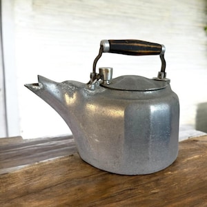 May include: A vintage aluminum tea kettle with a dark wooden handle and a spout. The kettle is sitting on a wooden surface. The kettle has a lid and a handle that swivels up and down.