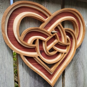 May include: A layered wooden heart wall art piece featuring a complex, interwoven knot design. The object is constructed from alternating light and dark wood tones, creating a dimensional effect. A single metal screw is visible in the center.