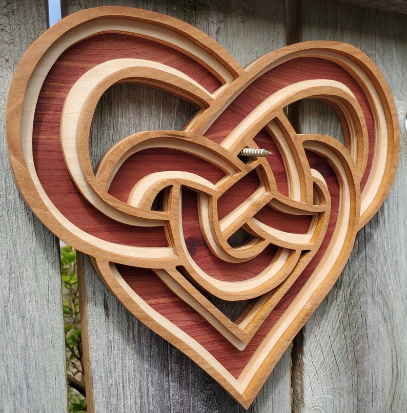 May include: A layered wooden heart wall art piece featuring a complex, interwoven knot design. The object is constructed from alternating light and dark wood tones, creating a dimensional effect. A single metal screw is visible in the center.