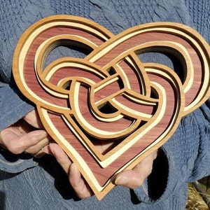 May include: A layered wooden heart sculpture featuring an intricate, interwoven knot design. The piece is constructed from alternating layers of deep reddish-brown wood and light cream-colored wood, creating a dimensional, multi-level effect.