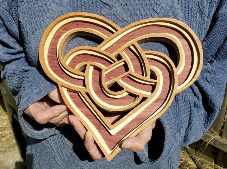May include: A layered wooden heart sculpture featuring an intricate, interwoven knot design. The piece is constructed from alternating layers of deep reddish-brown wood and light cream-colored wood, creating a dimensional, multi-level effect.