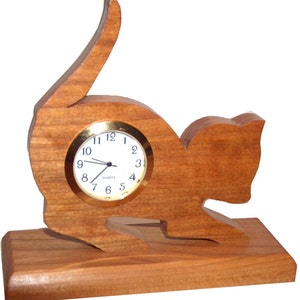 May include: A wooden clock shaped like a cat sitting on a wooden base. The clock face is white with black hands and gold numbers.
