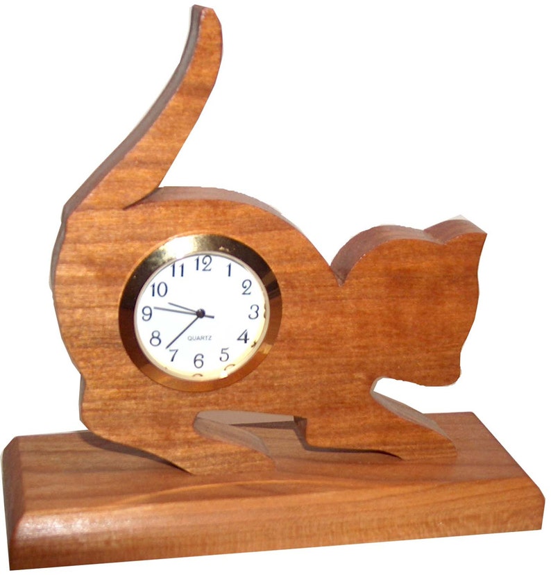 May include: A wooden clock shaped like a cat sitting on a wooden base. The clock face is white with black hands and gold numbers.