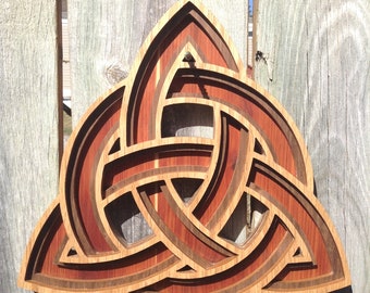 May include: A wooden Celtic knot, also known as a Trinity knot, is intricately carved with multiple layers of wood. The knot is a traditional symbol of love, faith, and hope. The knot is a light brown color with darker brown accents.
