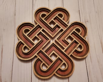 May include: A multi-layered wooden wall art piece featuring a symmetrical, interlocking geometric knot design. The object is constructed from stacked layers of wood in varying shades of light tan, medium brown, and dark reddish-brown tones.