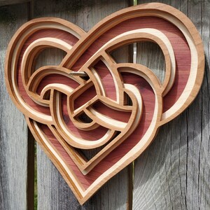May include: A heart-shaped wooden wall art piece featuring a layered, three-dimensional Celtic knot design. The object is constructed from alternating light and dark wood tones, showcasing a complex, interwoven pattern with a visible metal screw.