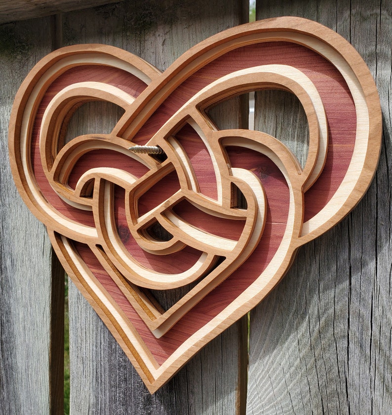May include: A heart-shaped wooden wall art piece featuring a layered, three-dimensional Celtic knot design. The object is constructed from alternating light and dark wood tones, showcasing a complex, interwoven pattern with a visible metal screw.