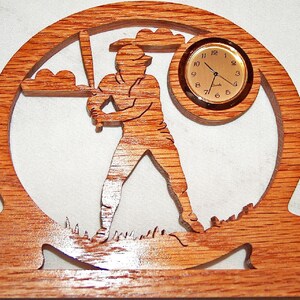 May include: Wooden baseball-themed clock with a quartz movement. The clock features a silhouette of a baseball player in a batting stance, a baseball, and a round clock face. The clock is made of light brown wood.