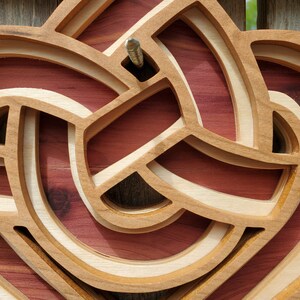 May include: A layered wooden wall art piece featuring an intricate, continuous knot design. The structure consists of light-colored, smooth-finished wooden bands arranged in an overlapping pattern, set against a deep reddish-brown wooden backing.