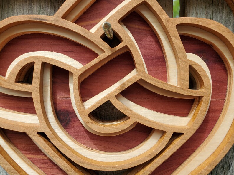 May include: A layered wooden wall art piece featuring an intricate, continuous knot design. The structure consists of light-colored, smooth-finished wooden bands arranged in an overlapping pattern, set against a deep reddish-brown wooden backing.