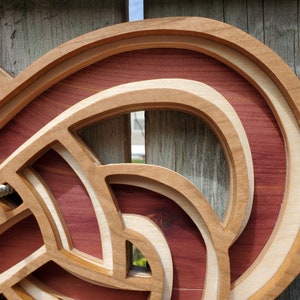 May include: A multi-layered wooden wall art piece featuring intricate, interlocking curved shapes. The design consists of alternating layers of light-toned wood and deep reddish-brown wood, creating a dimensional, stacked effect with visible depth.