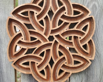 May include: A circular wooden wall art piece featuring a multi-layered, intricate Celtic knot design. The object is constructed from stacked wood panels with varying light and dark brown tones, creating a deep, dimensional, and geometric pattern.