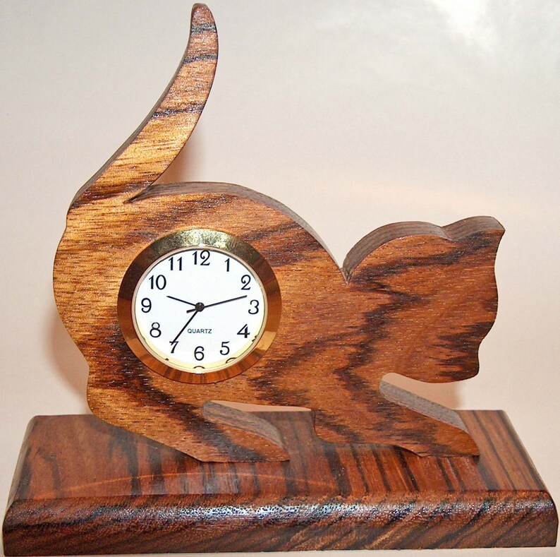 May include: A wooden cat clock with a brown wood grain finish. The clock has a round face with black numbers and hands. The clock is sitting on a wooden base.