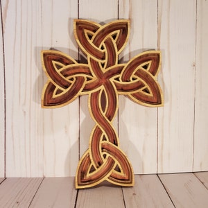 May include: A layered wooden cross featuring an intricate Celtic knot design. The object is constructed from multiple stacked layers of wood, showcasing alternating light tan and deep reddish-brown tones with visible wood grain throughout the structure.