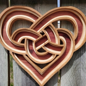 May include: A wooden heart-shaped wall decor with a Celtic knot design. The knot is intricately carved and features a layered effect with alternating shades of red and natural wood.