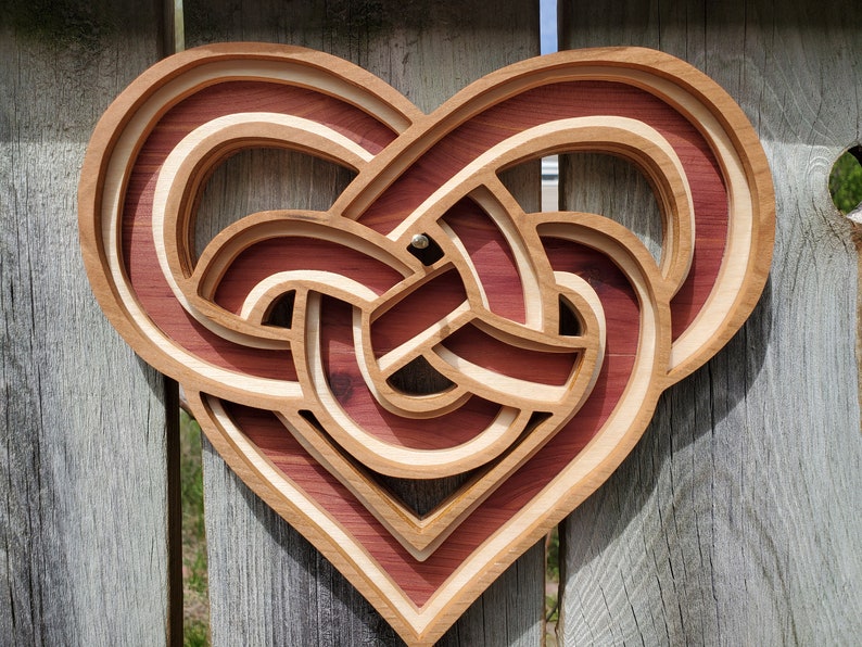 May include: A layered wooden wall art piece shaped like a heart with an intricate, interwoven knot design. The object features alternating light and dark wood tones, creating a three-dimensional effect with recessed channels and raised edges.