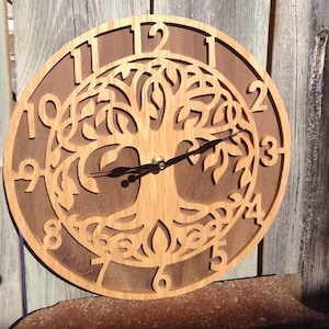 May include: A circular wall clock featuring a light-colored wood cutout of a tree of life design layered over a dark brown wood backing. The clock face displays large, cut-out numerals from one to twelve and has two thin black metal clock hands.