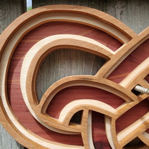 May include: A layered wooden wall art piece featuring an intricate, continuous knot design. The structure is composed of alternating light-colored wood and deep reddish-brown wood segments, creating a three-dimensional effect with a metal screw visible.